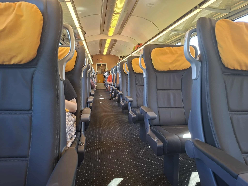 Wnętrze nowoczesnego wagonu pociągu z pustymi rzędami wygodnych siedzeń z żółtymi zagłówkami, rozciągającymi się wzdłuż korytarza, delikatnie oświetlone światłem dziennym wpadającym przez okna.