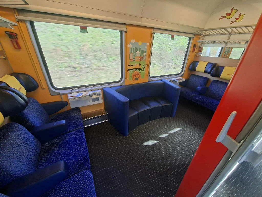 Przestronne wnętrze przedziału w pociągu z wygodnymi fotelami, miękką niebieską kanapą oraz dużymi oknami po bokach. Na ścianach znajdują się kolorowe elementy dekoracyjne, półka na bagaż, a przy oknie rozkładany stolik.