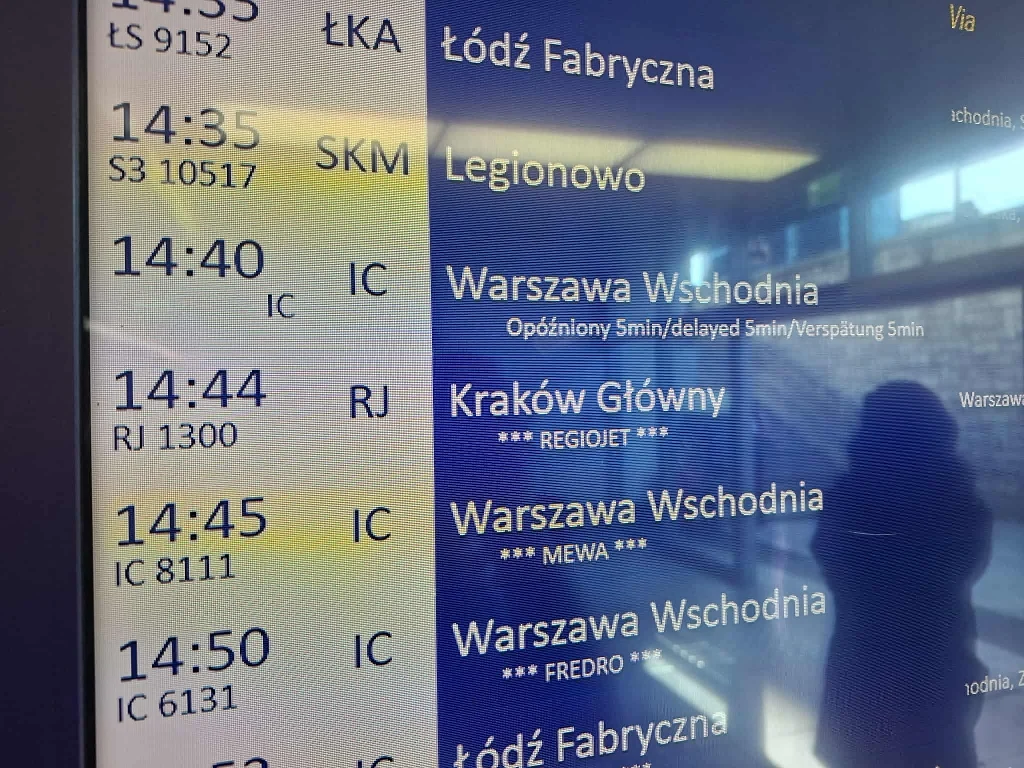 Tablica elektroniczna z rozkładem jazdy pociągów na dworcu kolejowym, widoczne godziny odjazdów, kierunki podróży do różnych miast, numery pociągów oraz sylwetka osoby odbijająca się w ekranie.