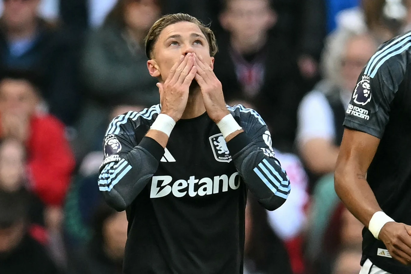 Piłkarz ubrany w czarną koszulkę z logo Betano wykonuje gest skierowania rąk do ust, prawdopodobnie w geście emocjonalnym, na tle rozmytej publiczności stadionu.