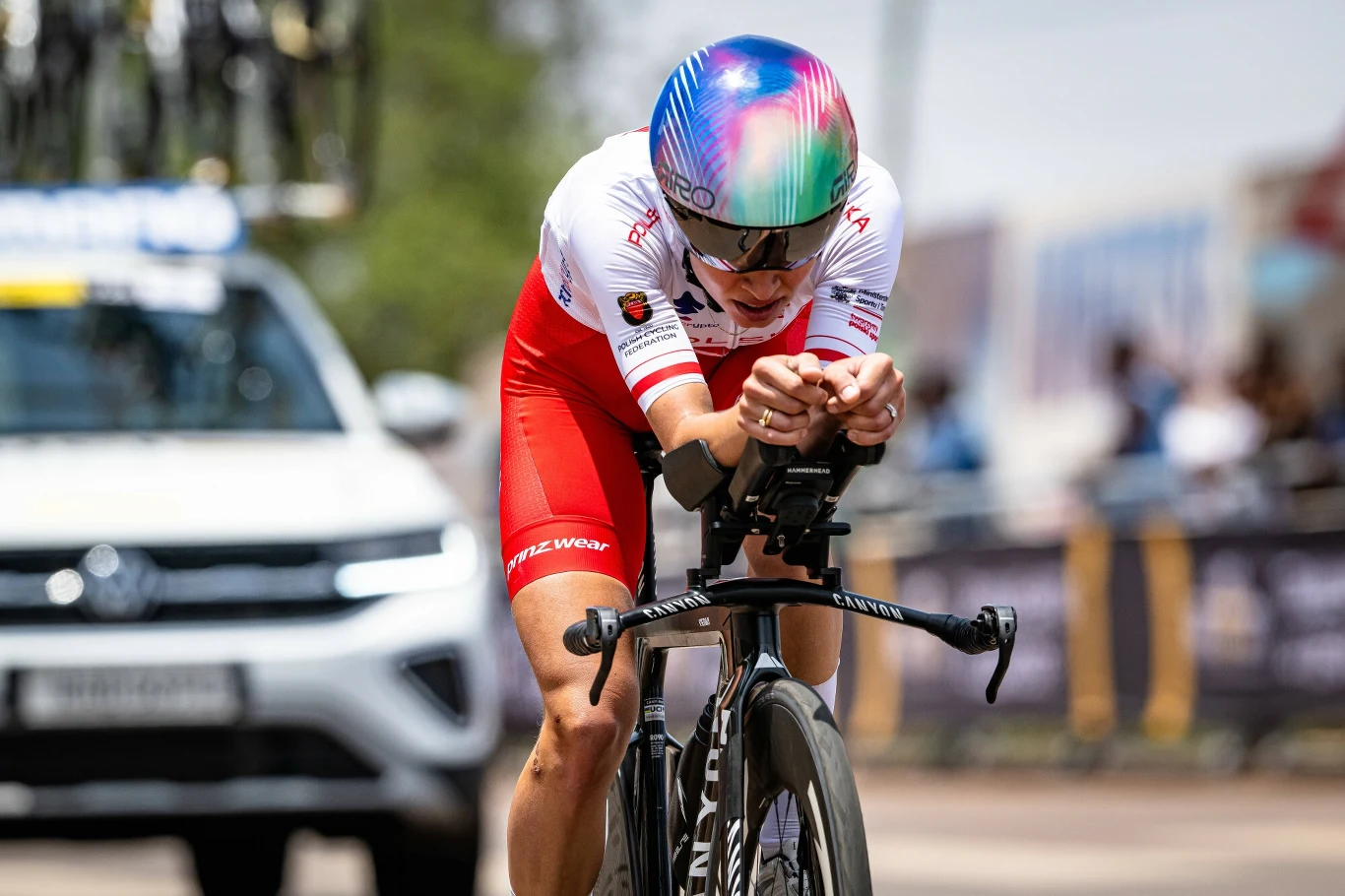 Kolarz ubrany w biało-czerwony strój sportowy oraz aerodynamiczny kask jedzie na rowerze szosowym podczas wyścigu ulicznego, na drugim planie porusza się samochód z rowerami na dachu.