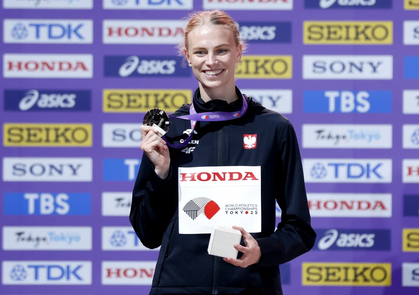 Uśmiechnięta sportsmenka w stroju reprezentacji Polski trzyma srebrny medal oraz dyplom, na tle ściany z logotypami sponsorów i nazwą wydarzenia sportowego.