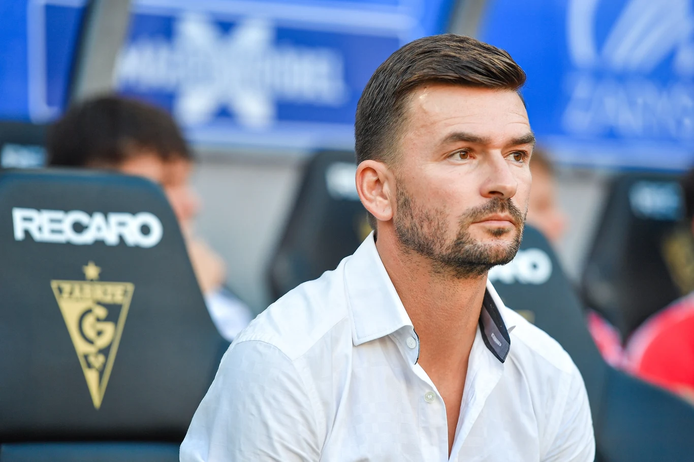 Michal Gasparik, trener nowego lidera Ekstraklasy Mężczyzna o krótkich, ciemnych włosach i zadbanym zaroście siedzi na stadionowej ławce rezerwowych, ubrany w białą koszulę, w tle widoczne rozmyte postacie oraz napisy na oparciach siedzeń.