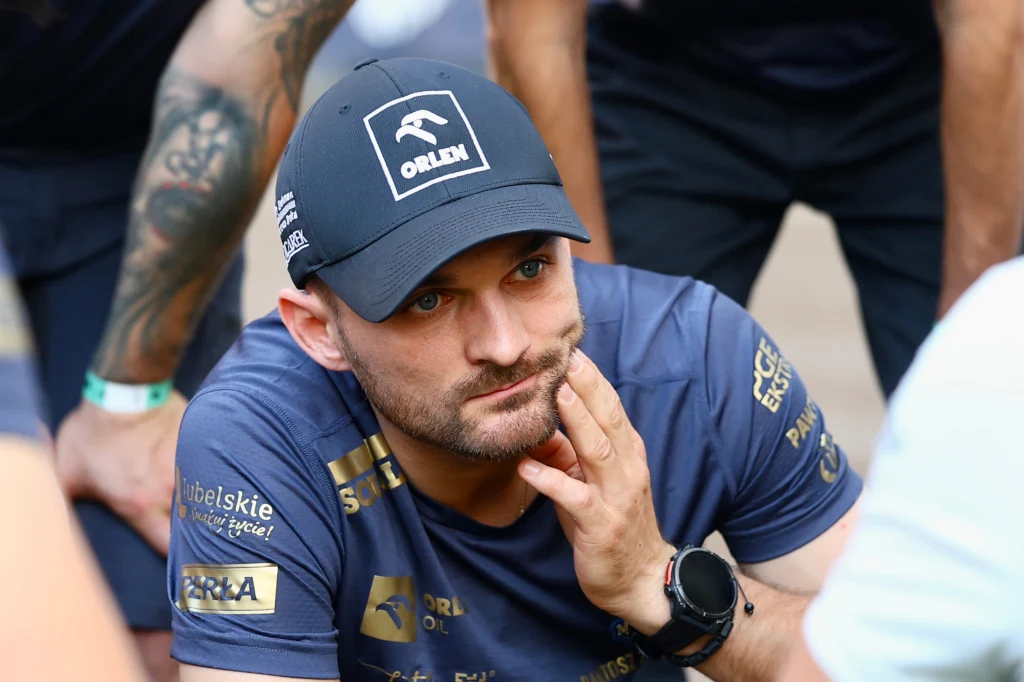 Mężczyzna w sportowej czapce z logo oraz granatowej koszulce oparty na dłoni, otoczony przez osoby w sportowych ubraniach, skupiona twarz i widoczne tatuaże na rękach w tle.