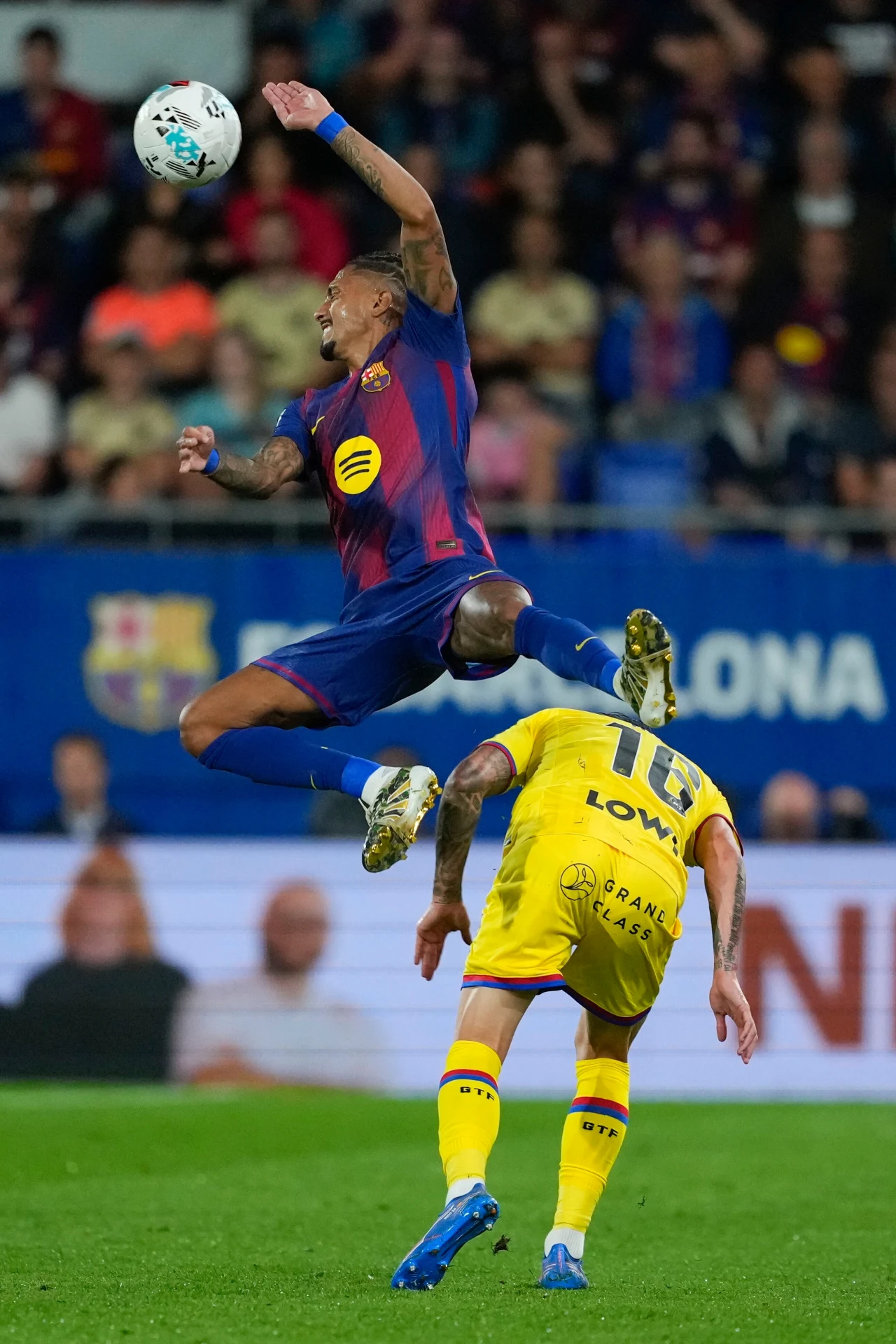 Piłkarz w barwach klubu FC Barcelona wyskakuje wysoko nad przeciwnikiem w żółtym stroju podczas walki o piłkę w powietrzu na tle widowni stadionu.
