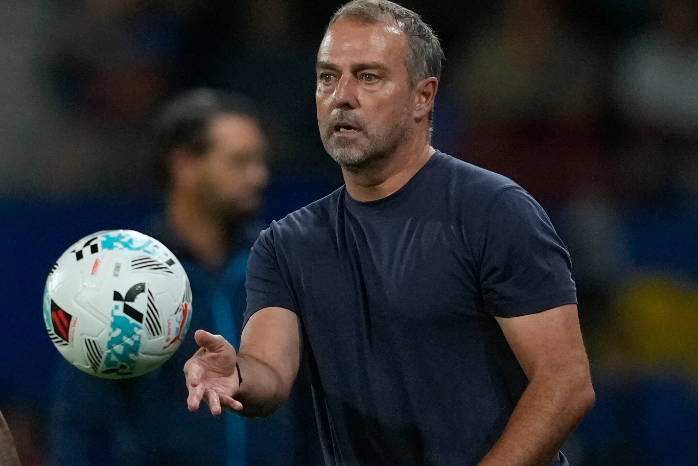 Mężczyzna ubrany w ciemny t-shirt próbuje złapać piłkę do piłki nożnej, w tle widoczna rozmyta postać oraz niewyraźne elementy stadionu.