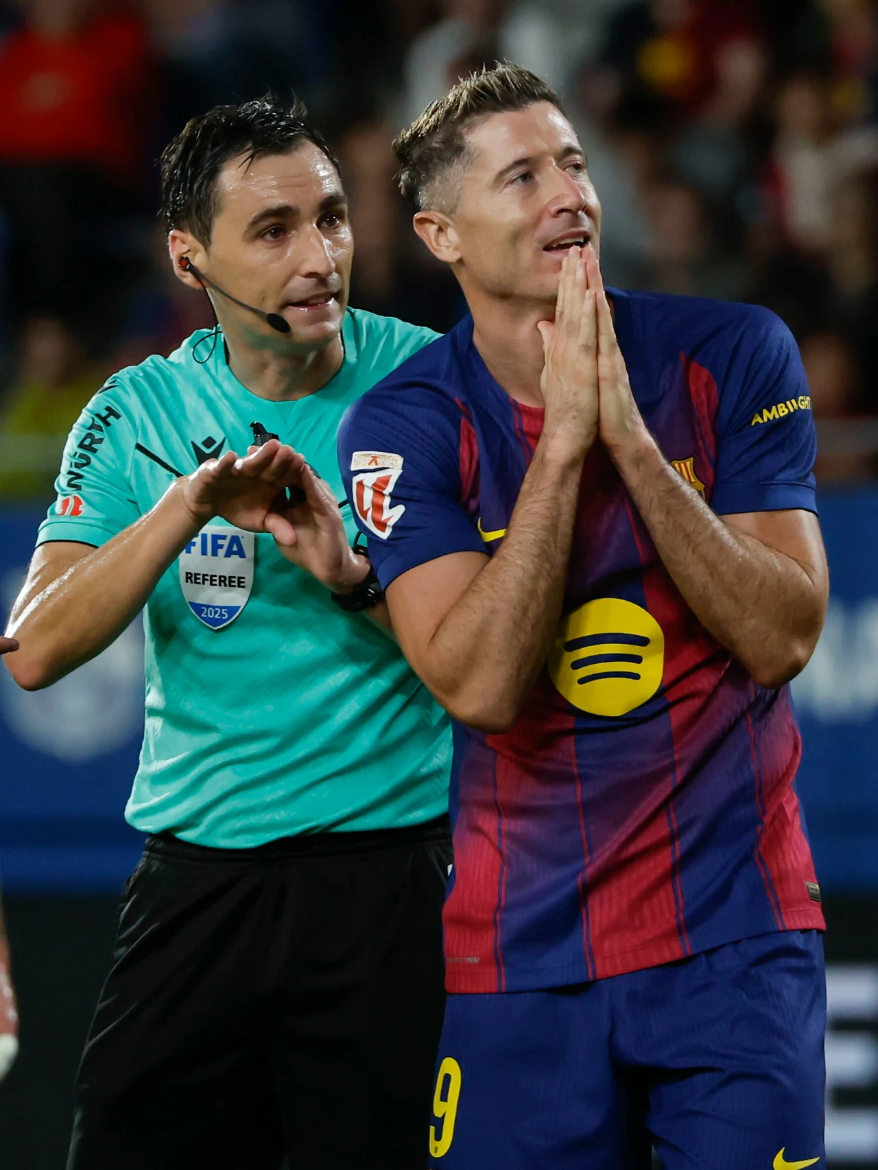 Robert Lewandowski w trakcie meczu z Getafe CF Piłkarz w koszulce FC Barcelony gestykuluje z rękami złożonymi przy twarzy, obok niego stoi sędzia w stroju z logo FIFA, który wykonuje gest ręką, tło zamazane, dominuje sportowa atmosfera.