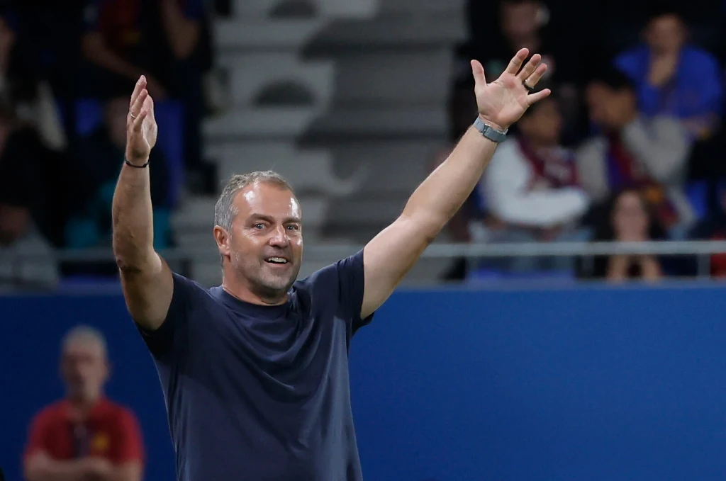 Po niedzielnym triumfie trener Hansi Flick nie krył entuzjazmu Uśmiechnięty mężczyzna w ciemnej koszulce stoi z uniesionymi rękami na tle stadionu, wokół widownia i osoby w tle.