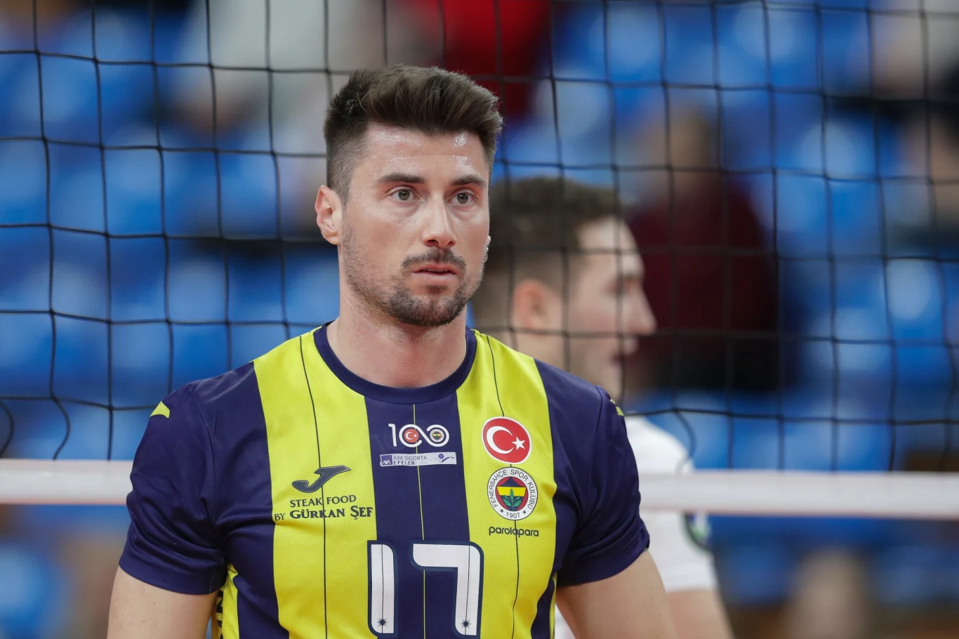 Murat Yenipazar Zawodnik siatkarski w żółto-granatowej koszulce z numerem 17 stoi przed siatką na hali sportowej, w tle niewyraźny sylwetka innego sportowca i rozmyte niebieskie trybuny.