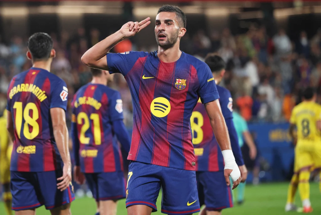 Ferran Torres Piłkarz FC Barcelony wykonuje gest podczas meczu, wokół kilku zawodników w strojach drużyny, w tle widoczni rywale ubrane na żółto oraz rozmyta publiczność na trybunach.