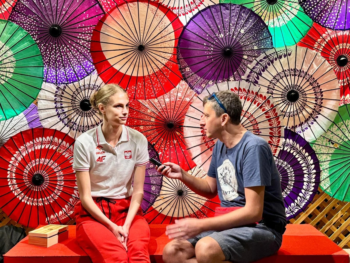 Rozmowa dwóch osób siedzących na czerwonej ławce, w tle wielokolorowe japońskie parasole tworzące efektowną dekorację; jedna osoba trzyma mikrofon, druga ubrana w strój reprezentacyjny z emblematami Polski.