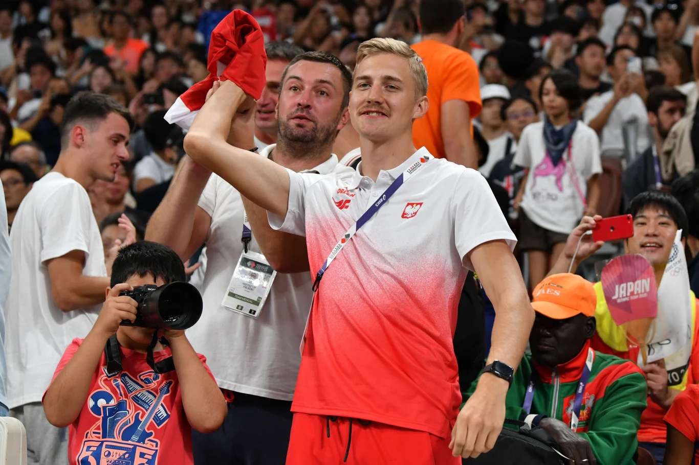 Uśmiechnięty sportowiec w stroju z logo Polski, trzymając czerwoną koszulkę, stoi wśród kibiców na trybunach, wokół tłum ludzi, fotograf robi zdjęcie, w tle widoczne jest logo Japonii.