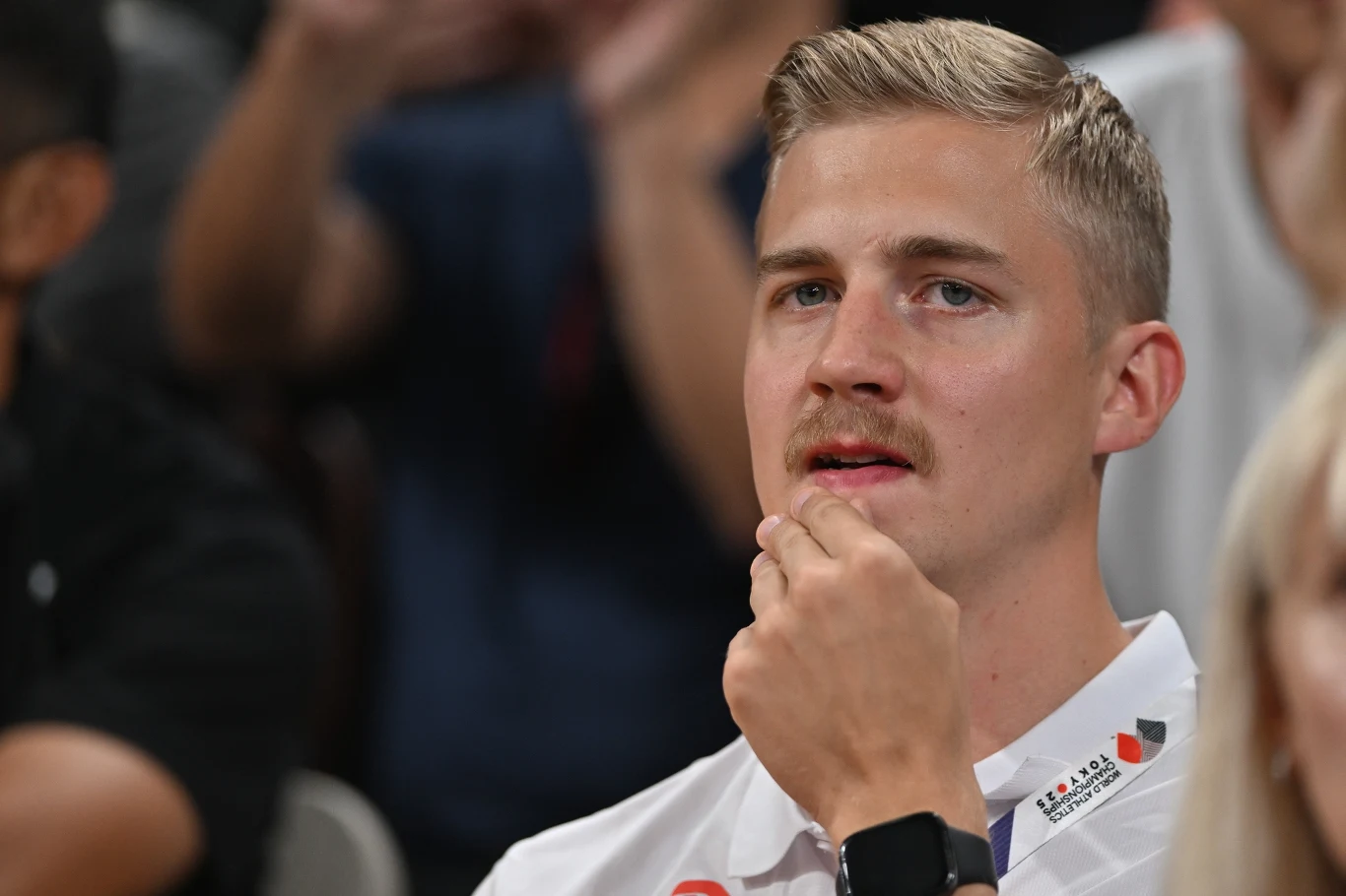 Mężczyzna o blond włosach i wąsach w białej koszulce z plakietką siedzi na widowni, podpiera brodę dłonią i patrzy w kierunku sceny, w tle rozmyte sylwetki innych osób.
