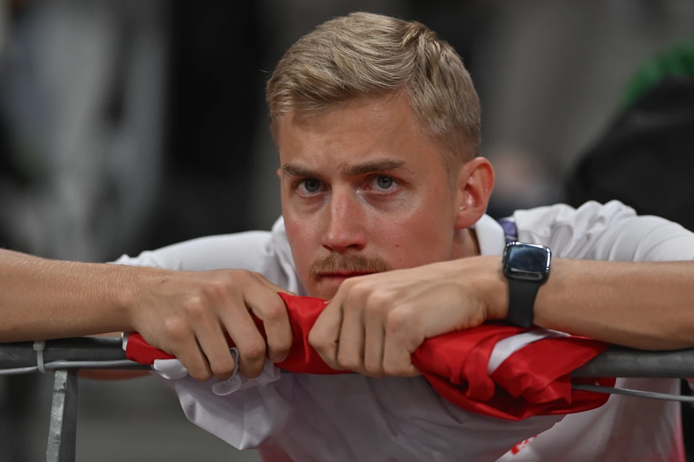 Młody mężczyzna o blond włosach z poważnym wyrazem twarzy oparty na barierce, trzymający w dłoniach zwiniętą biało-czerwoną flagę.