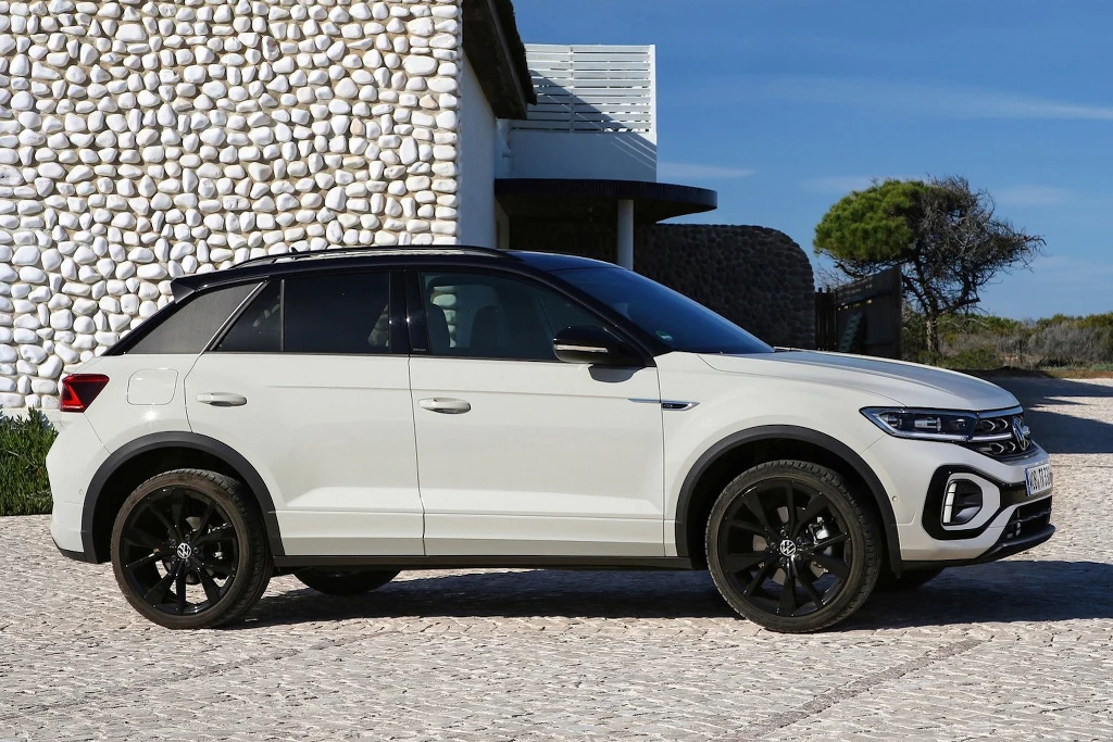 Volkswagen T-Roc to nowy król europejskich dróg. W sierpniu był najlepiej sprzedającym się nowym autem w Europie Biały samochód osobowy typu SUV zaparkowany na brukowanym podjeździe przed nowoczesnym budynkiem o białej elewacji z ozdobnymi kamieniami, w tle widoczne drzewo i fragment nieba.