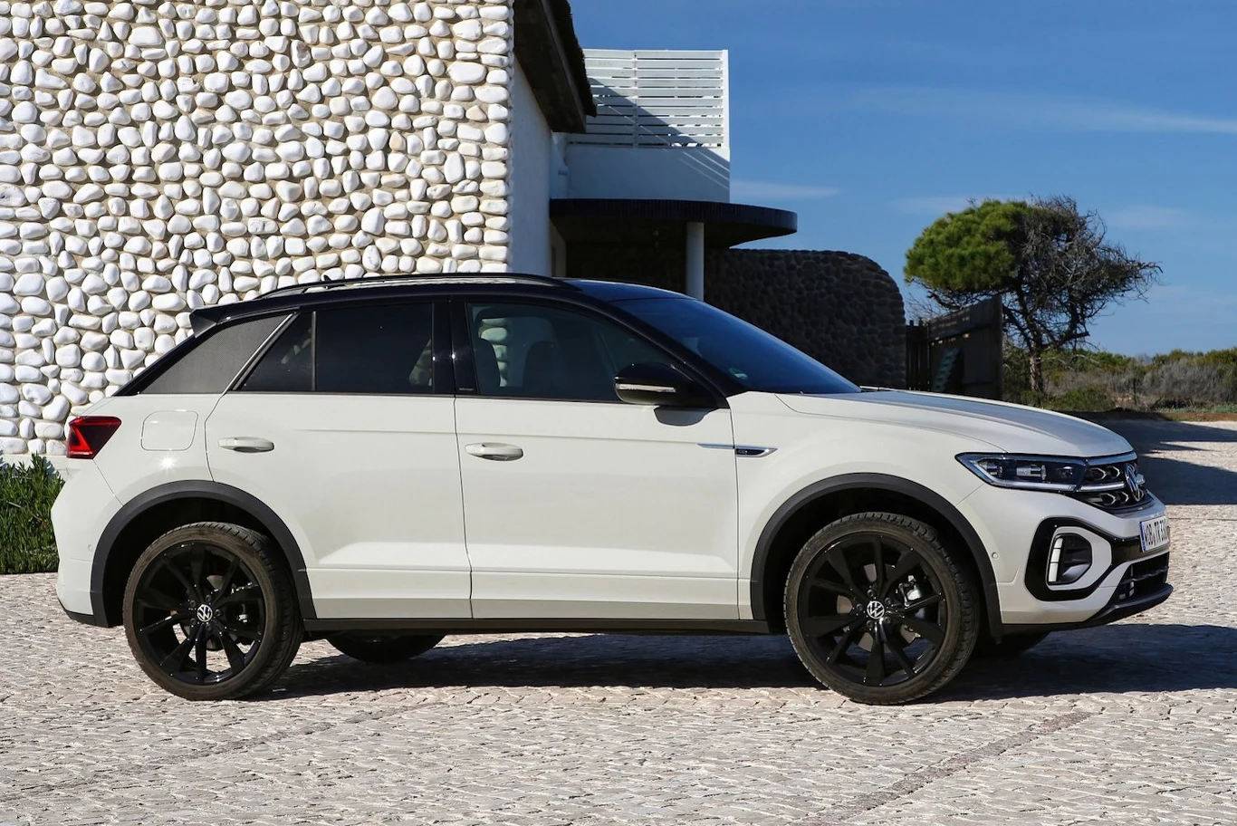 Biały samochód osobowy typu SUV zaparkowany na brukowanym podjeździe przed nowoczesnym budynkiem o białej elewacji z ozdobnymi kamieniami, w tle widoczne drzewo i fragment nieba.