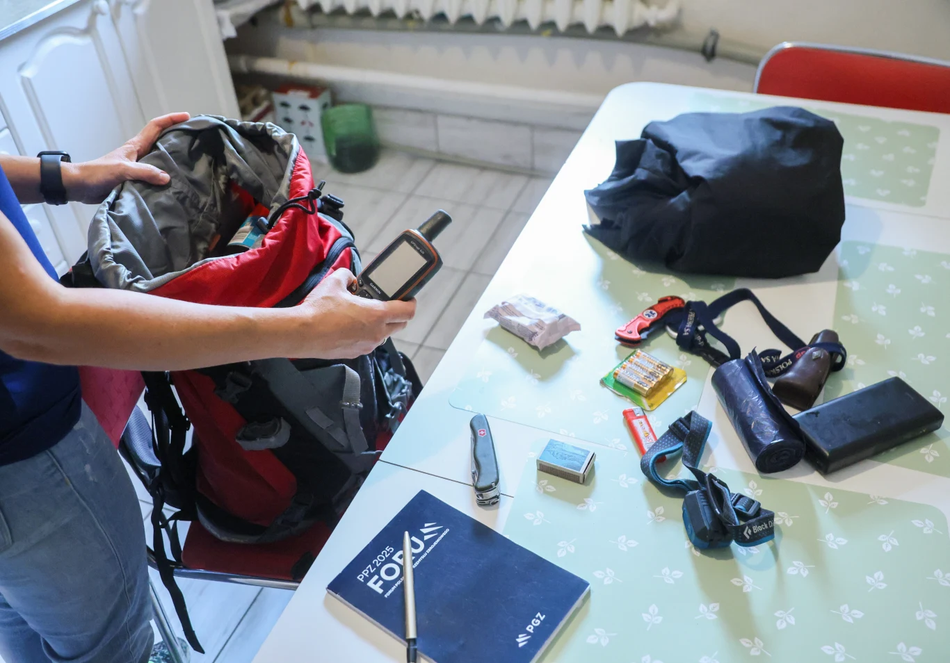 Plecak ewakuacyjny. Jaki plecak wybrać i co do niego spakować? Sprzęt turystyczny i rzeczy osobiste rozłożone na stole, w tym plecak, notes, nożyk, czołówka, lornetka, aparat, mapnik oraz jedzenie, osoba pakująca plecak przed wyprawą.
