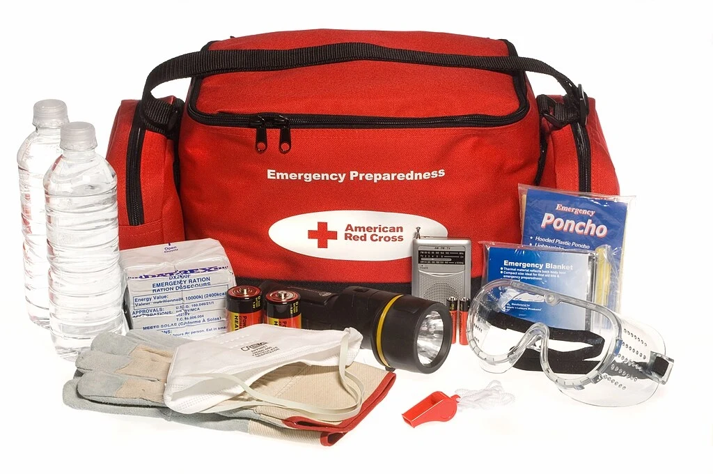 Czerwony worek z napisem Emergency Preparedness, należący do Amerykańskiego Czerwonego Krzyża, otoczony zestawem przedmiotów ratunkowych takich jak butelki wody, rękawiczki, latarka, baterie, okulary ochronne, radio, gwizdek, koc termiczny, poncho i ma...