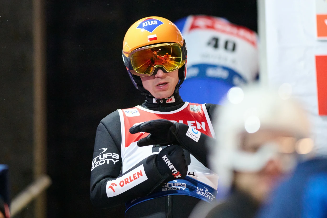 Zawodnik w stroju do skoków narciarskich stoi przygotowany do startu, ubrane gogle i kask, na piersi widoczne logo sponsorów oraz numer startowy, w tle rozmazany sylwetka innego uczestnika zawodów.