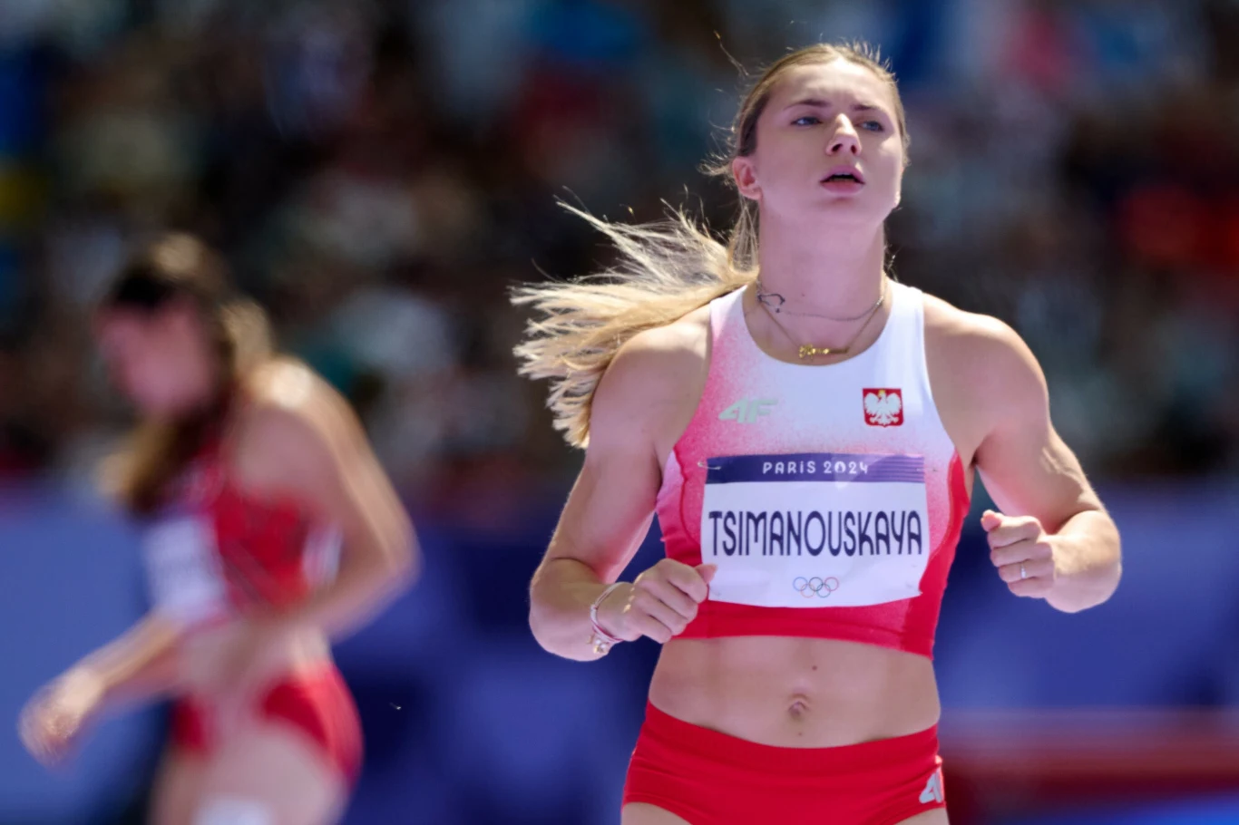 Zawodniczka w stroju sportowym z nazwiskiem na koszulce bierze udział w zawodach lekkoatletycznych, w tle widoczna jest rywalka i rozmyte trybuny z widownią.
