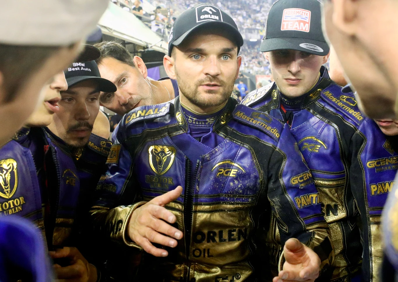 Grupa żużlowców ubranych w kolorowe kombinezony teamowe, stojących w gęstym kręgu, skupionych na rozmowie, otoczonych przez ludzi i reflektory stadionu.