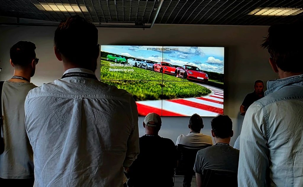 Grupa osób uczestnicząca w prezentacji motoryzacyjnej, gdzie na dużym ekranie wyświetlona jest grafika z samochodami Porsche na torze wyścigowym oraz hasłem dotyczącym techniki jazdy i bezpieczeństwa na torze, sala o industrialnym wystroju, uczestnicy ...