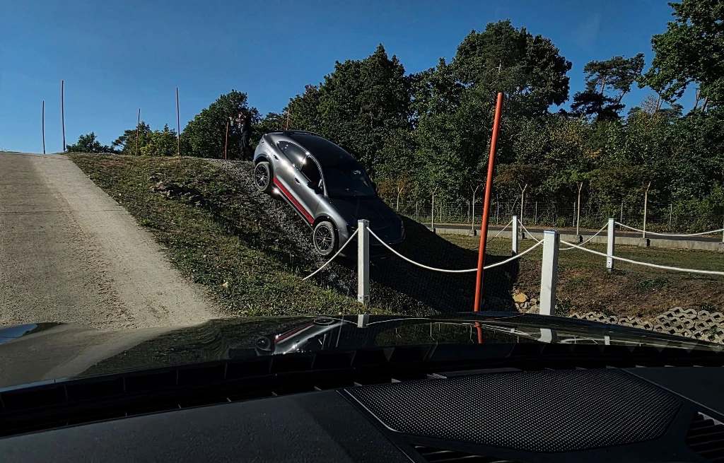 Samochód terenowy pokonuje strome wzniesienie na specjalnie przygotowanym torze testowym, na pierwszym planie fragment kokpitu innego auta, w tle drzewa i wyznaczone bariery.