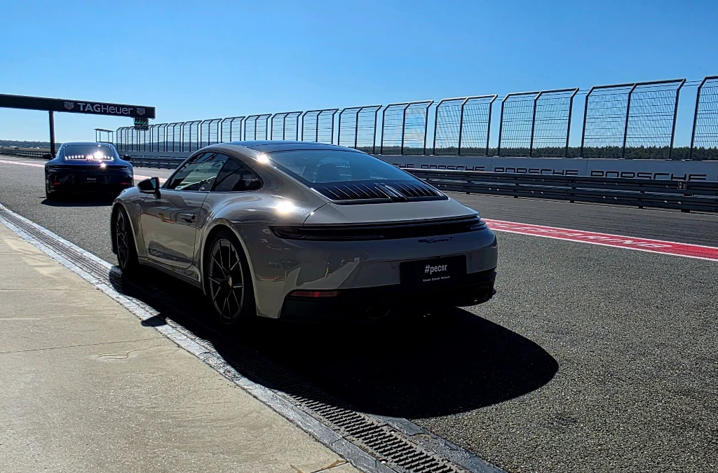 Jasnoszary sportowy samochód marki Porsche znajduje się na torze wyścigowym przy bandzie, w tle drugi samochód nadjeżdżający po torze, otoczenie zabezpieczone barierkami ochronnymi, nawierzchnia asfaltowa lśniąca od słońca.