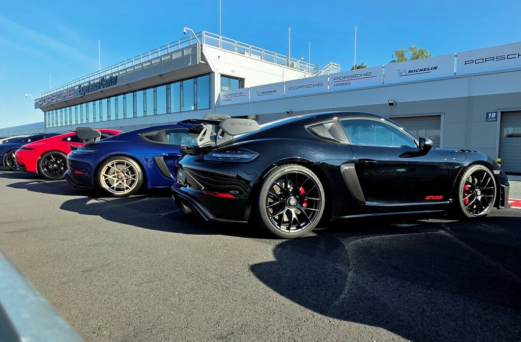 Rzędy sportowych samochodów Porsche ustawione pod budynkiem centrum wyścigowego, wyraźnie widoczne modele w czarnym, niebieskim i czerwonym kolorze, nowoczesna architektura zaplecza oraz czyste niebo w tle.