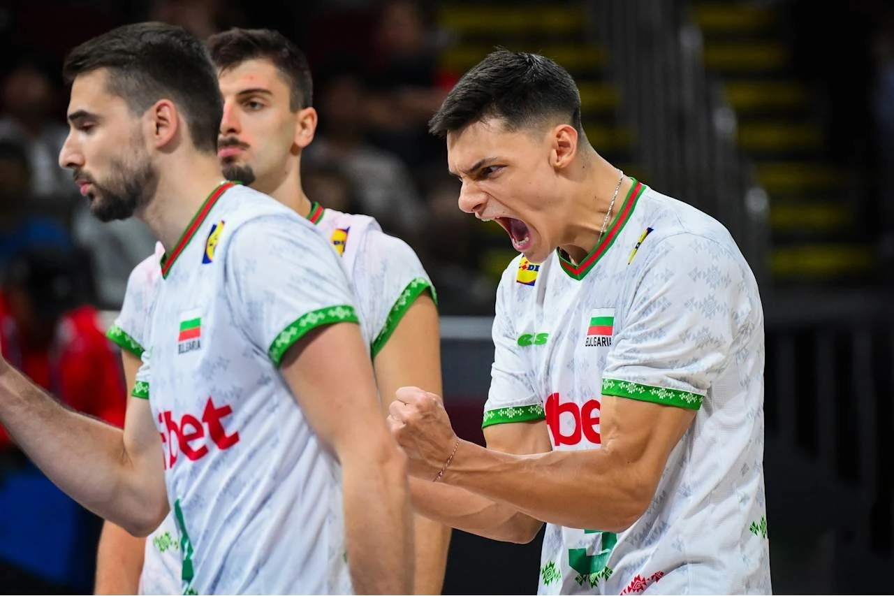 Zawodnik drużyny siatkarskiej w białej koszulce z zielonymi akcentami wyraża silne emocje po udanej akcji, zaciskając pięść i krzycząc, obok niego stoją jego koledzy z drużyny odwróceni lekko bokiem