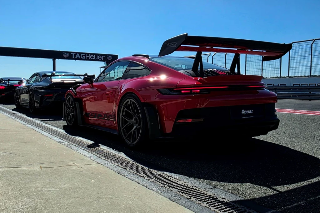Czerwony sportowy samochód Porsche z dużym tylnym spojlerem stoi zaparkowany na torze wyścigowym obok innego auta, w tle widoczne barierki i oznaczenie TAG Heuer.