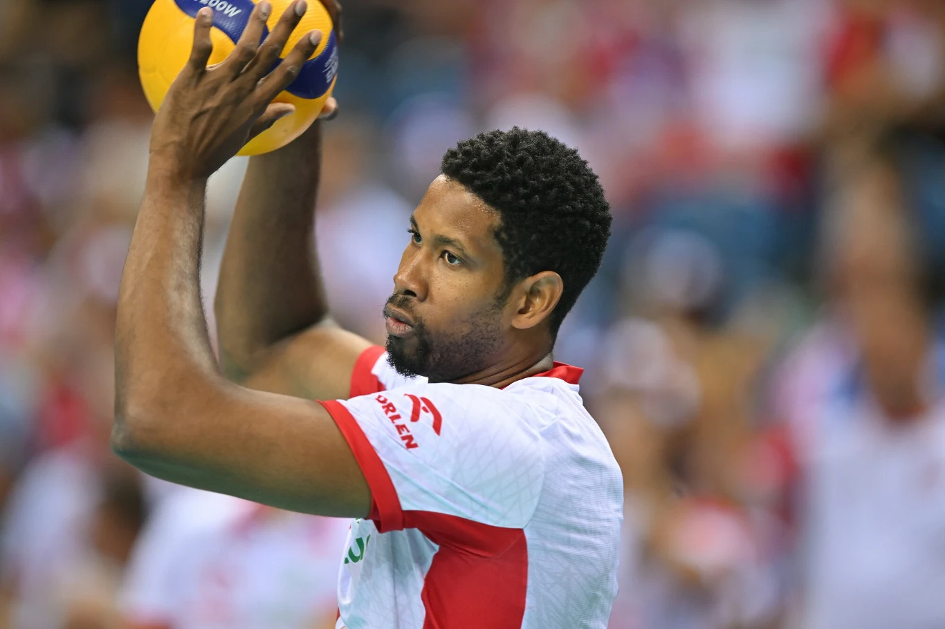 Siatkarz trzymający piłkę do siatkówki w gotowości do rozegrania akcji, ubrany w biało-czerwony strój sportowy, w tle widoczna rozmyta publiczność.