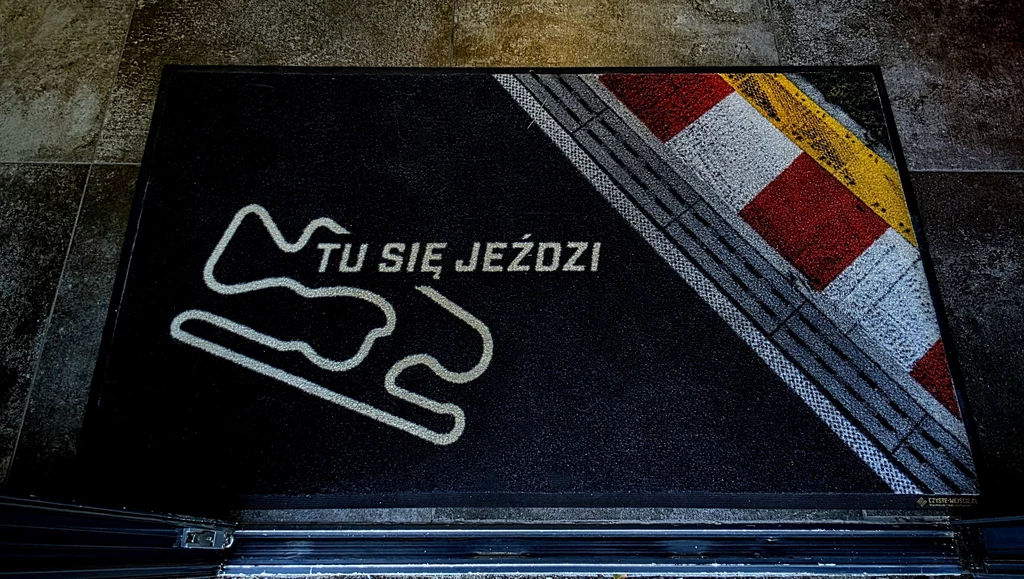 Dywanik z napisem 'TU SIĘ JEŹDZI', widoczny schemat fragmentu toru wyścigowego oraz pasy namalowane na asfalcie z żółtym i czerwonymi elementami, całość tworzy motyw związany z wyścigami.