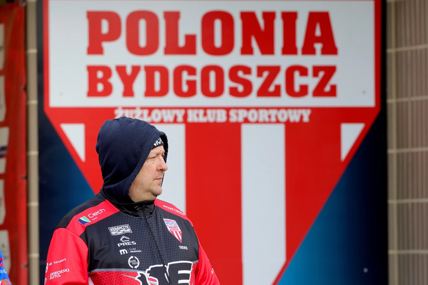 Mężczyzna w czarnej czapce i sportowej kurtce z logo drużyny żużlowej stoi przed dużym szyldem Polonia Bydgoszcz, żużlowego klubu sportowego.
