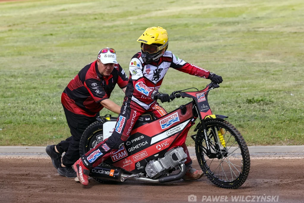 Zawodnik żużlowy w żółtym kasku siedzi na motocyklu na torze, a mężczyzna ubrany w czerwono-czarną odzież techniczną pomaga mu ruszyć pchając motocykl, w tle widoczny jest zielony murawowy fragment toru.
