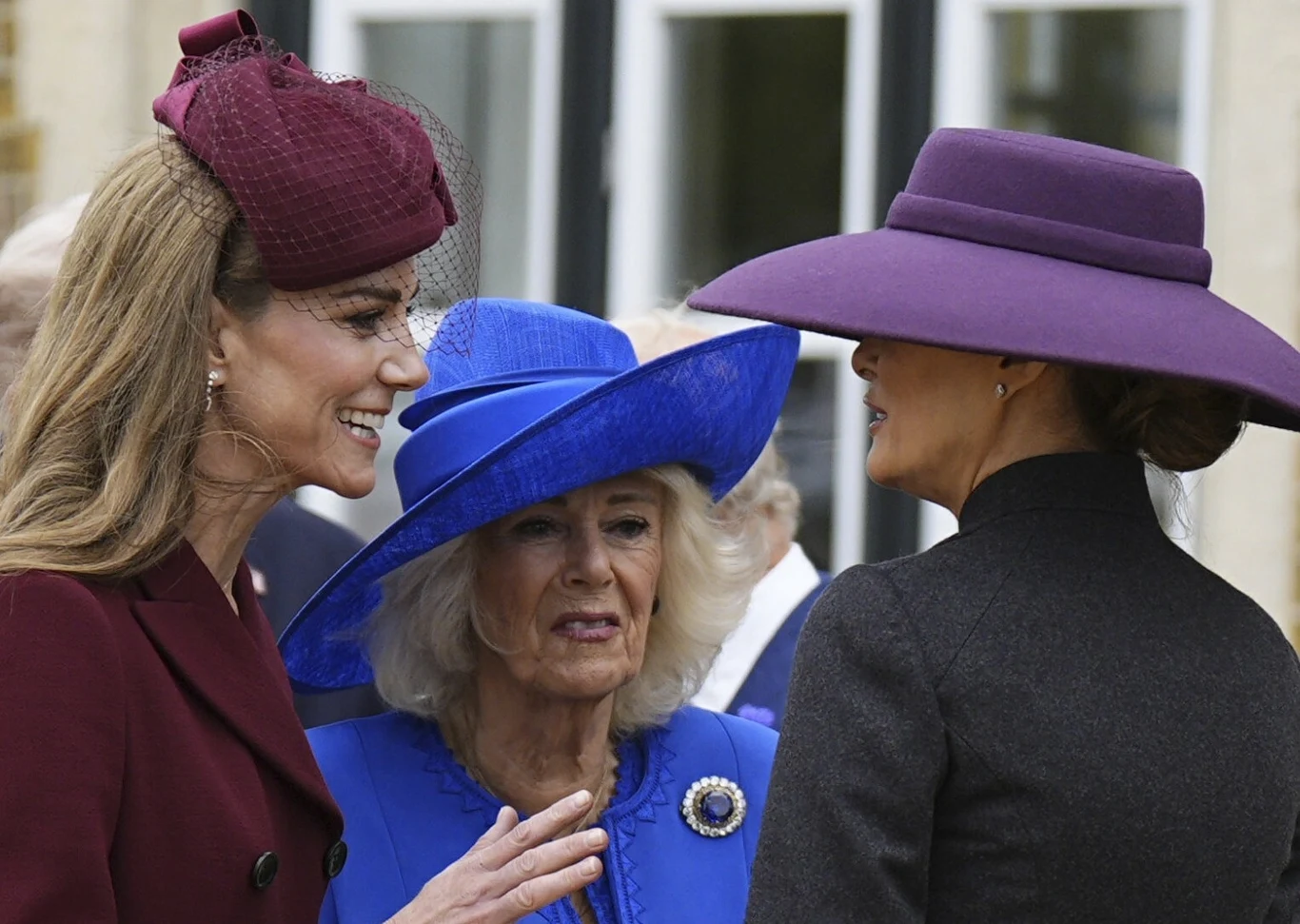 Księżna Kate, królowa Kamila i Melania Trump w 2025 r. Trzy elegancko ubrane kobiety rozmawiają ze sobą na zewnątrz, dwie w kapeluszach w intensywnych kolorach — bordo i niebieskim — oraz jedna w dużym kapeluszu w kolorze fioletowym, wszystkie ubrane w stylowe płaszcze.