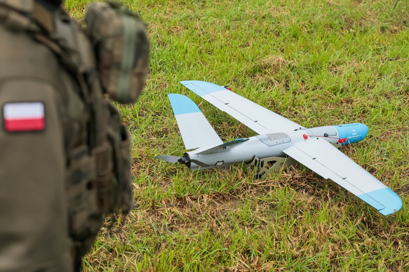 Polska rozpocznie budowę własnego systemu antydronowego. Zdj. ilustracyjne Dron o stałych skrzydłach ustawiony na trawie, obok fragment sylwetki osoby w mundurze z naszywką flagi Polski.