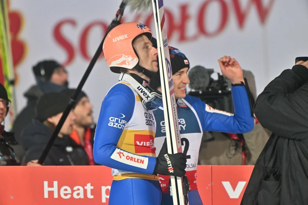 Dawid Kubacki i Kamil Stoch Zawodnik w kombinezonie do skoków narciarskich trzymający narty, stojący obok innego sportowca podczas zawodów, w tle widoczni kibice oraz baner reklamowy.