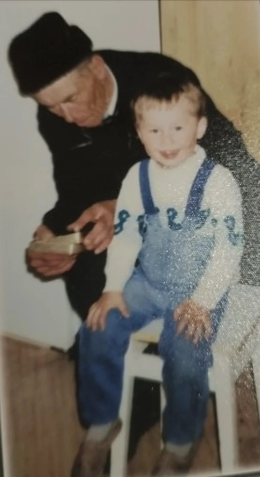 Starszy mężczyzna w czapce pochylony przy jasnowłosym dziecku siedzącym na stołku, oboje w pomieszczeniu o jasnych ścianach, mężczyzna trzyma niewielkie drewniane pudełko.