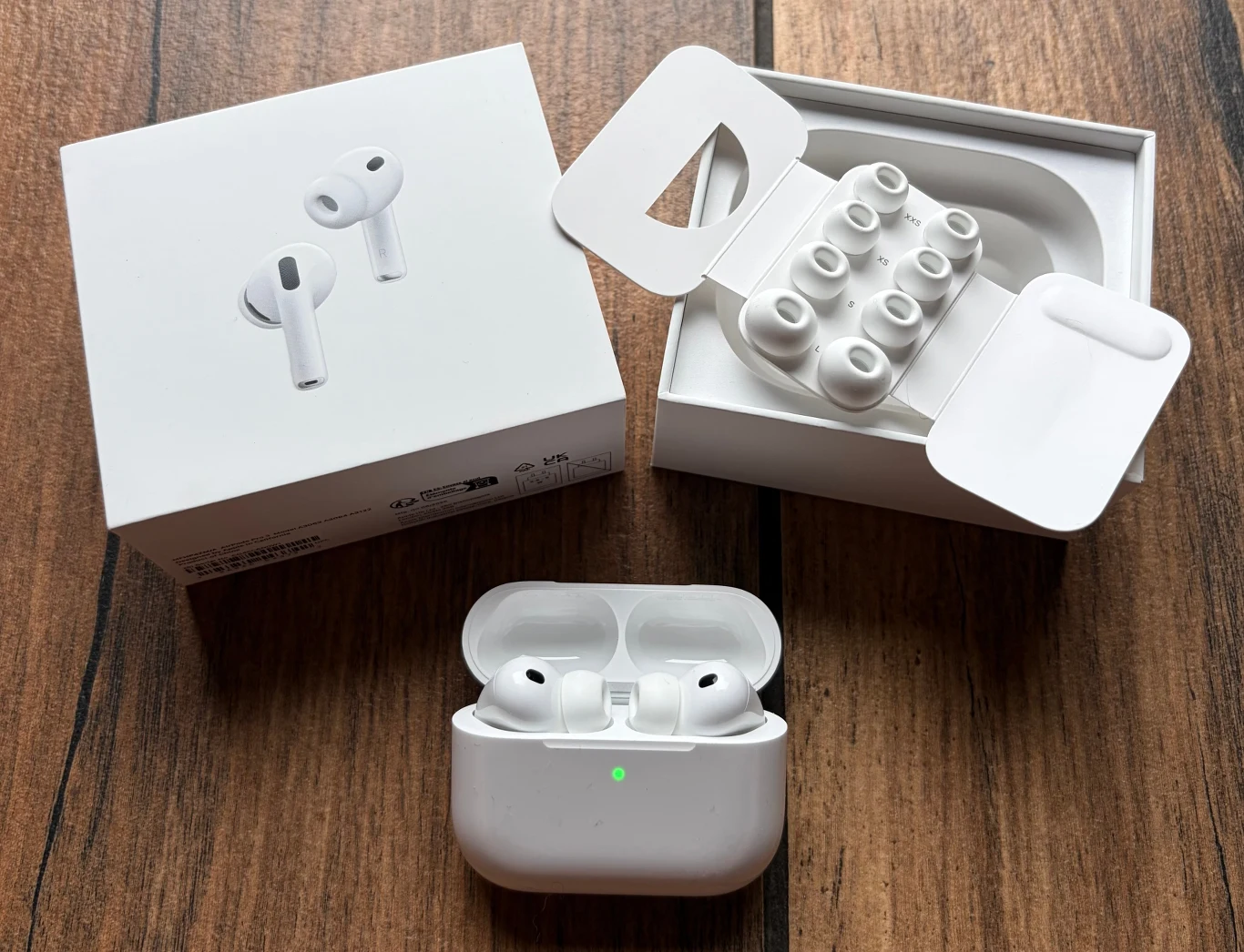 Zestaw bezprzewodowych słuchawek dousznych z białym etui ładującym, oryginalnym opakowaniem oraz wymiennymi silikonowymi końcówkami, umieszczony na drewnianej powierzchni.