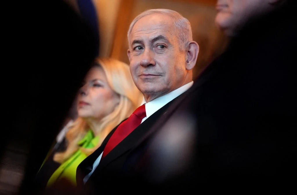 Mężczyzna w garniturze z czerwonym krawatem siedzi w dobrze oświetlonym pomieszczeniu obok kobiety z blond włosami, ubranej w żółte ubranie. Ujęcie skupia się na jego twarzy i wyrazie skupienia.