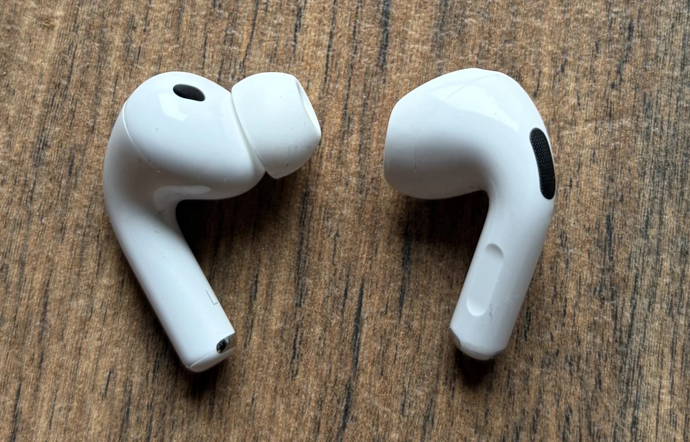 Dwie białe bezprzewodowe słuchawki douszne leżące na drewnianej powierzchni, ukazane z bliska, w orientacji równoległej do siebie.