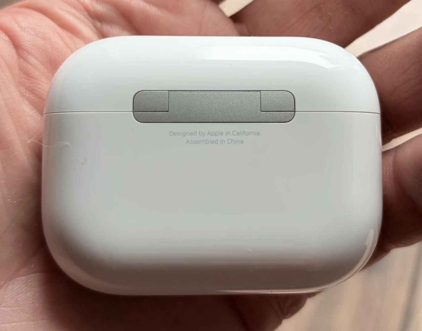 Białe etui na słuchawki bezprzewodowe z widocznym zawiasem oraz napisem informującym o zaprojektowaniu przez Apple w Kalifornii, trzymane w ludzkiej dłoni.