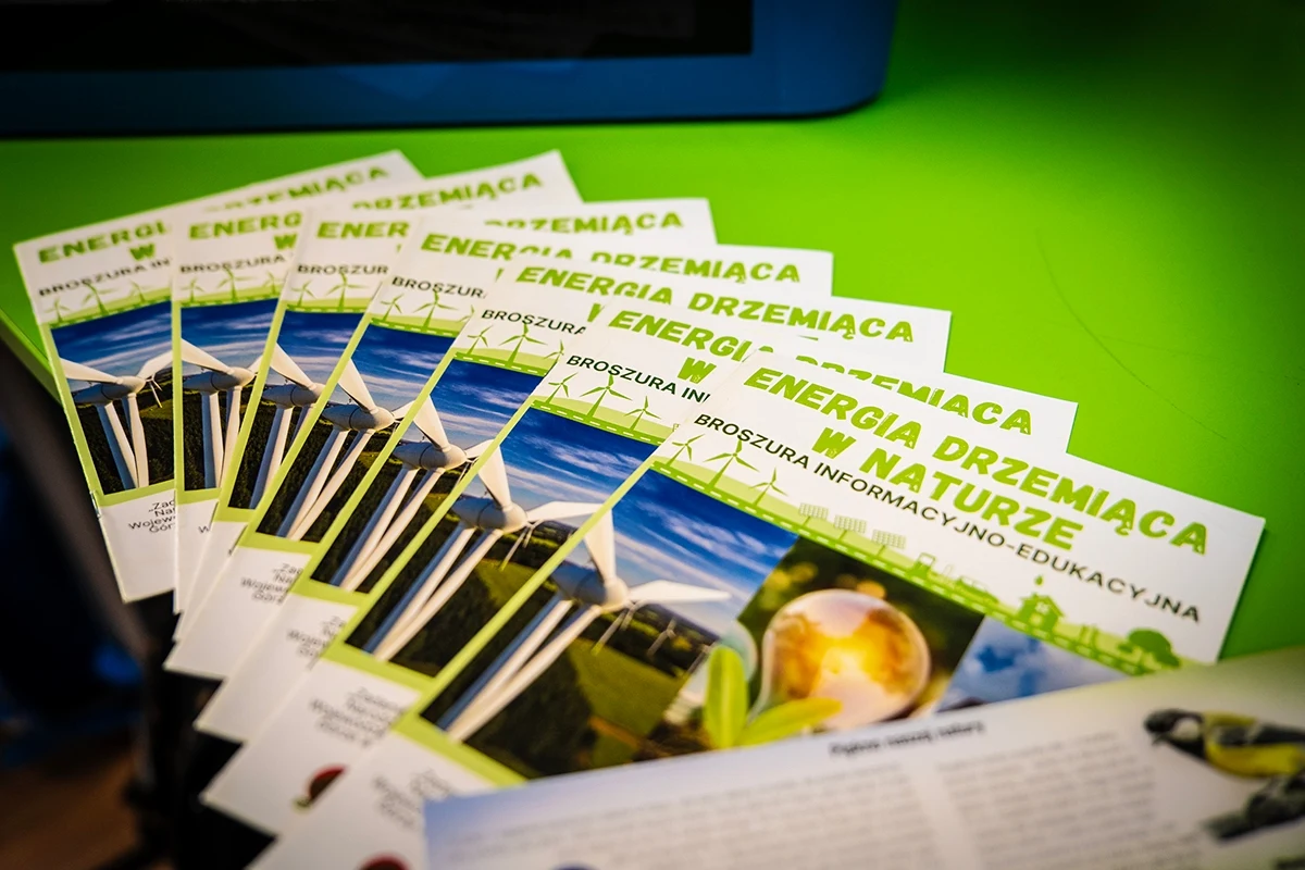 Stos materiałów edukacyjnych dotyczących energii odnawialnej, na których widoczne są ilustracje turbin wiatrowych oraz krajobrazu pełnego zieleni.