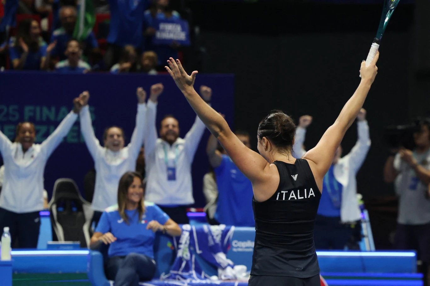 Tenisistka z uniesionymi rękami w geście triumfu na korcie, na plecach widoczny napis Italia; w tle drużyna ubrana na biało i niebiesko cieszy się energetycznie z sukcesu zawodniczki.