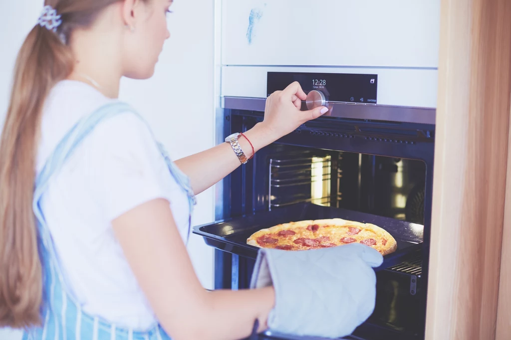 Młoda kobieta wkłada pizzę do piekarnika, używając rękawicy ochronnej, widoczny elektroniczny panel sterowania piekarnika oraz ciepłe światło wnętrza.