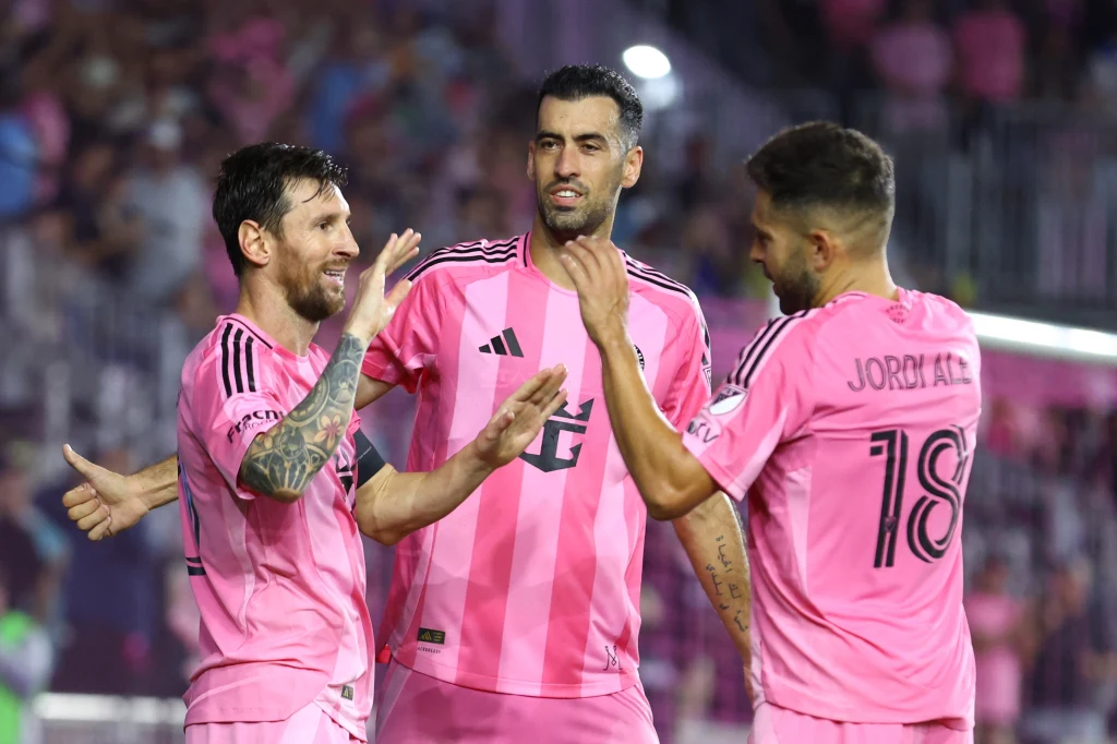 Lionel Messi, Sergio Busquets i Jordi Alba Trzech piłkarzy w różowych koszulkach drużyny piłkarskiej świętujących sukces na boisku, jeden z nich podnosi rękę do przybicia piątki, tłum kibiców w tle.