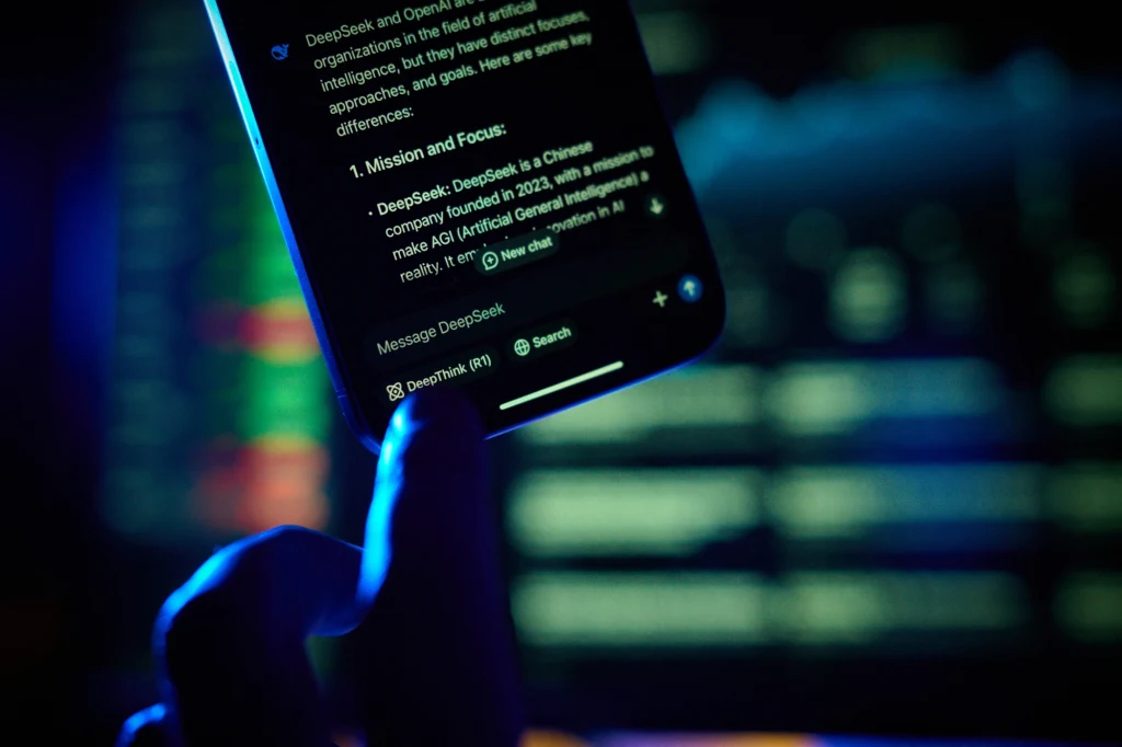 Sztuczna inteligencja odbiera pracę w wielu branżach. AI miała być tylko wsparciem, ale jest inaczej. Zdj. ilustracyjne Ręka trzyma smartfon z wyświetlonym tekstem na ekranie, w tle rozmazane kolorowe światła i ekran komputera, całość utrzymana w ciemnej, technologicznej kolorystyce.