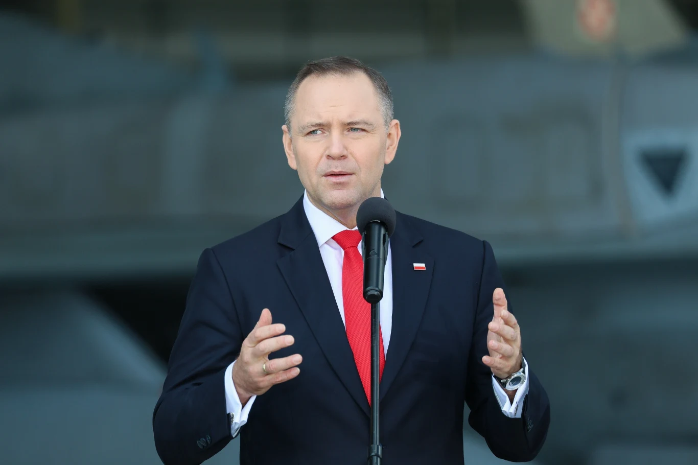 Prezydent Karol Nawrocki Mężczyzna w garniturze z czerwonym krawatem przemawia do mikrofonu na tle wojskowego sprzętu, przypięta do klapy marynarki flaga Polski podkreśla oficjalny charakter wystąpienia.