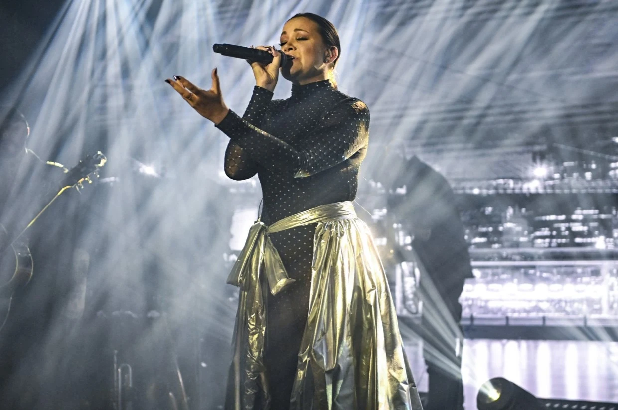 Dominika Gawęda jest wokalistką Blue Cafe Wokalistka śpiewająca na scenie, ubrana w oryginalny strój z połyskującą spódnicą, trzymająca mikrofon, w tle efektowna scenografia z jasnymi światłami oraz zabudową sceniczną.